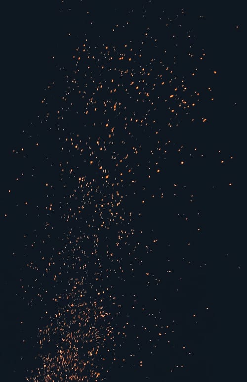 Orange, pixelish sparks rise up from a bonfire just out of the frame of the photo, which was taken at night.